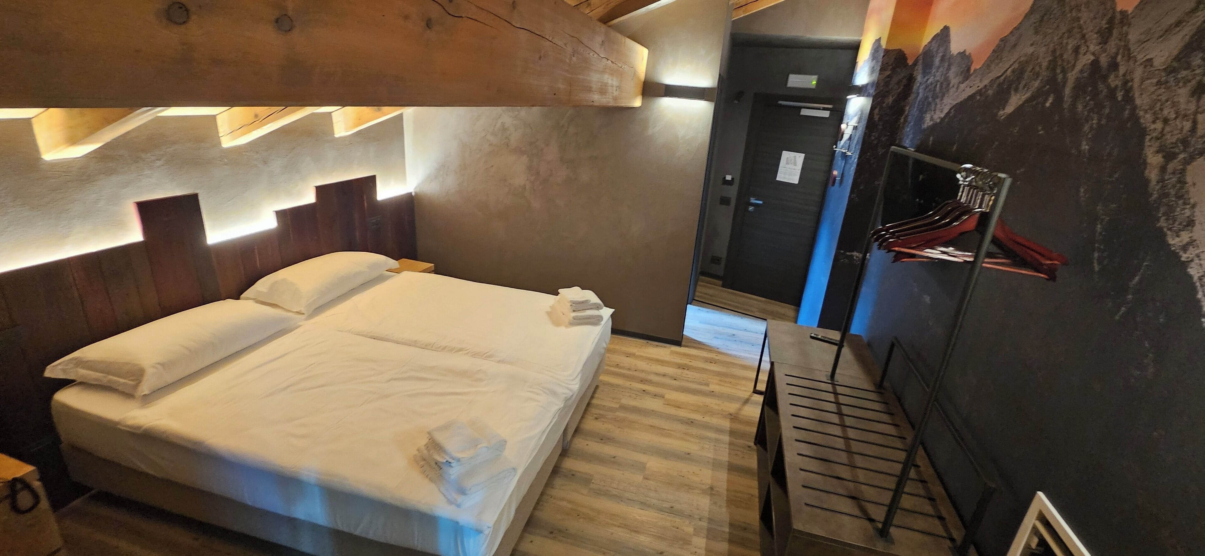 Habitación de hotel con una cama, cabecera de madera, perchero en la pared y una puerta al fondo.