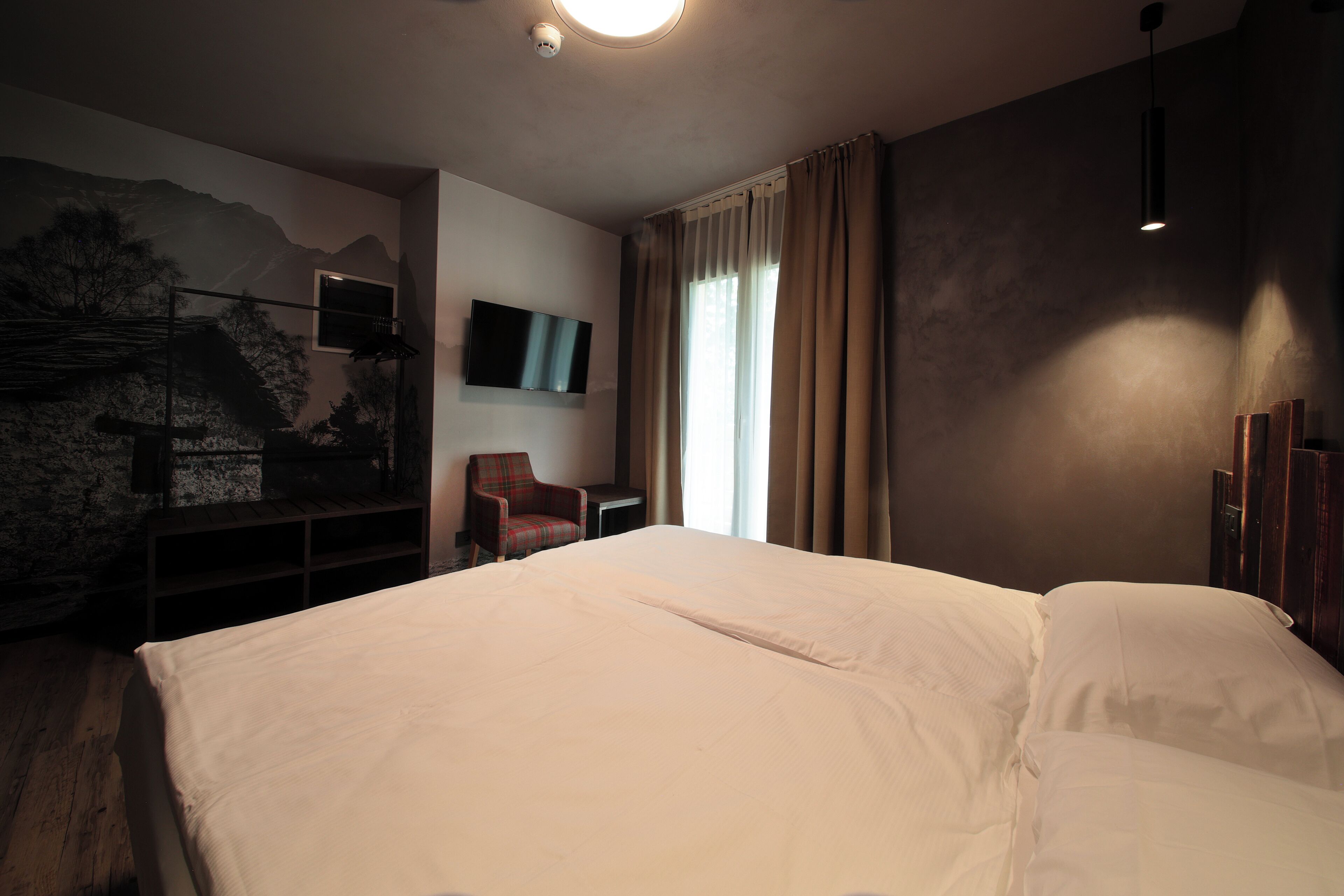 A hotel room with a large bed, a chair, a TV mounted on the wall, and a painting of a mountain scene.
