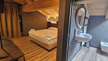A bedroom with a bed, bedside table, and a wooden ceiling. The bathroom includes a sink, mirror, and bidet.