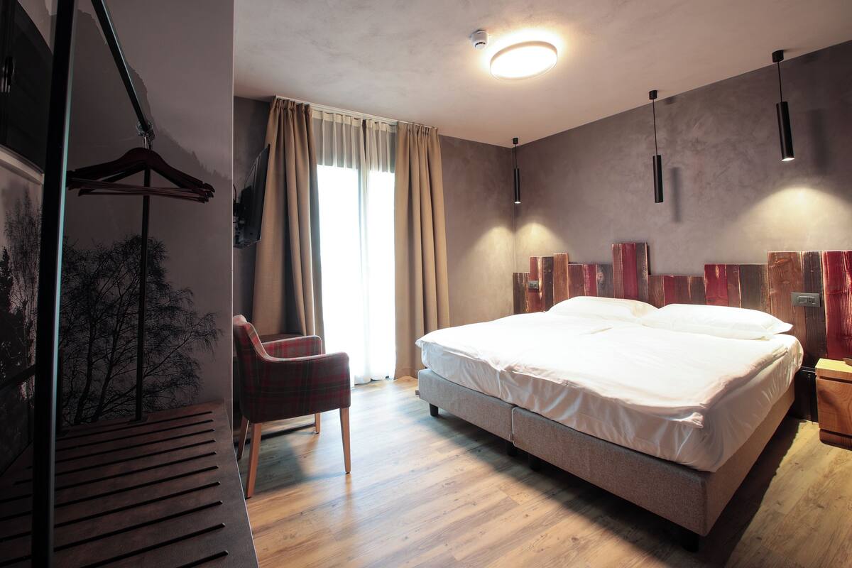 A modern bedroom with a large bed, a chair, a hanging coat rack, and a view of trees through a window.