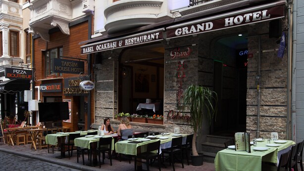 An outdoor dining area with tables set for two, a Grand Hotel sign, and a Taksim House Hotel sign.