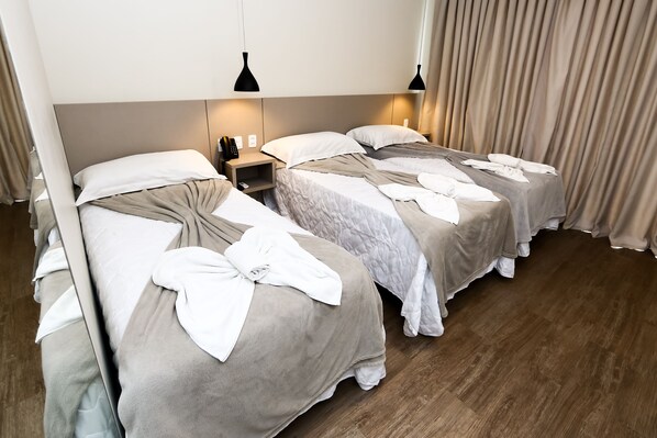 A hotel room with two beds, each with a white pillow and a towel. There are two bedside tables with lamps, and the room has wooden flooring.