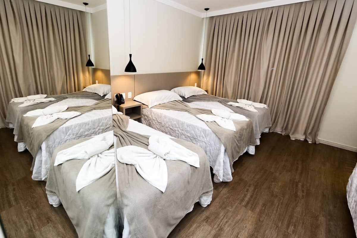 A hotel room with four beds arranged in a U-shape, each with white linens and a beige blanket. There are two black pendant lights above the beds, and the room has wooden flooring.