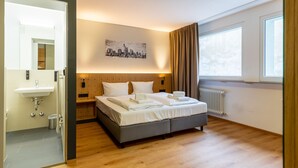 A modern hotel room with a large bed, a bathroom with a sink, and a cityscape painting on the wall. - mk | hotel frankfurt (Frankfurt)