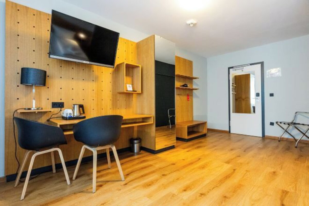A modern room with a wooden wall, a flat-screen TV, a desk with two chairs, a lamp, and a small kitchenette.