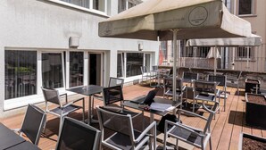 An outdoor patio with tables and chairs, a large umbrella, and a building with windows and balconies. - mk | hotel frankfurt (Frankfurt)