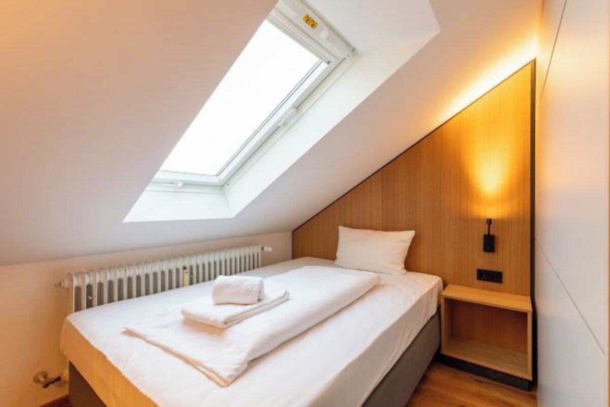 A modern bedroom with a skylight, a bed with white linens, a wooden headboard, and a bedside table with a lamp.