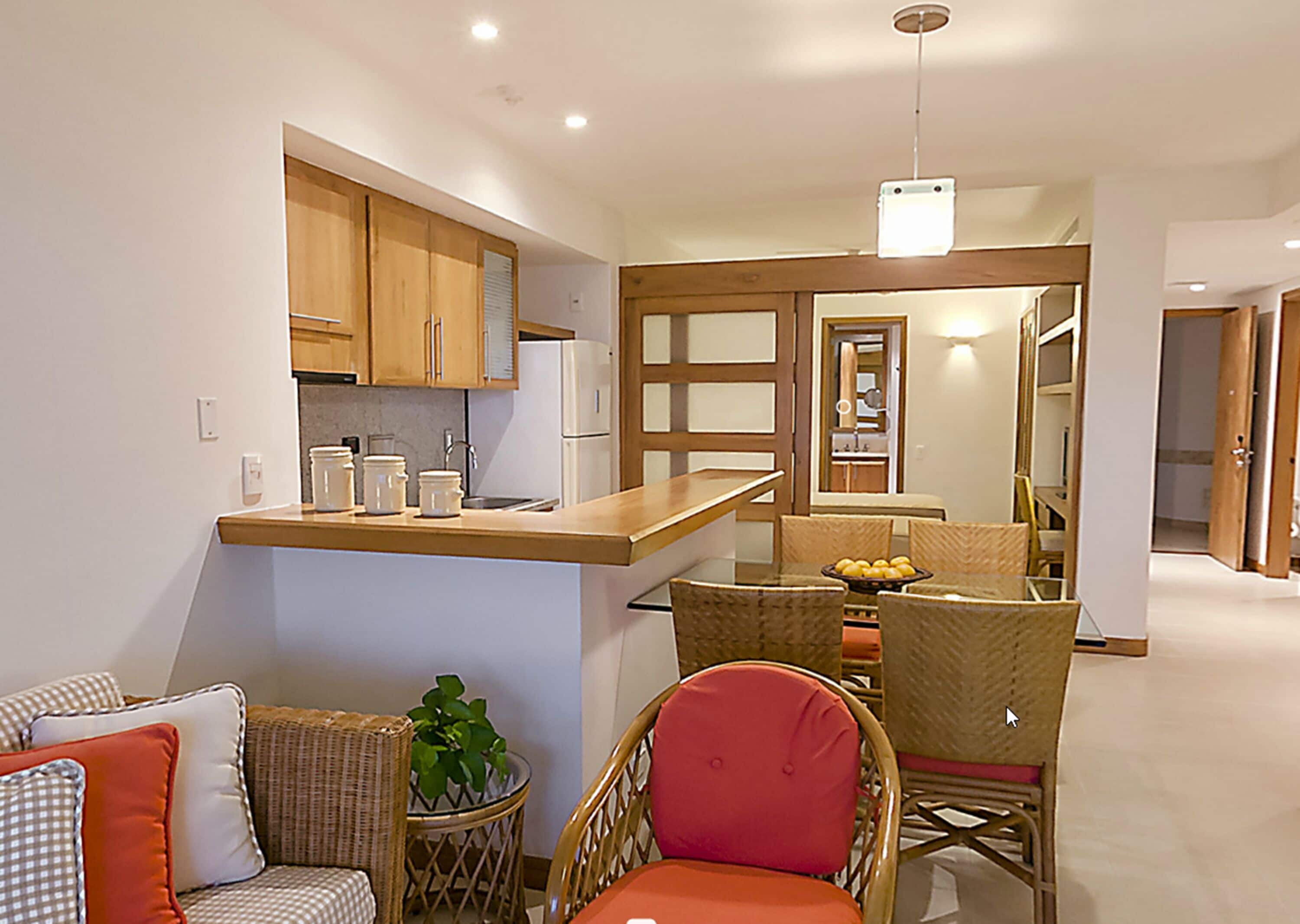A compact kitchen with wooden cabinets, a built-in fridge, and a dining area with wicker chairs and a table.