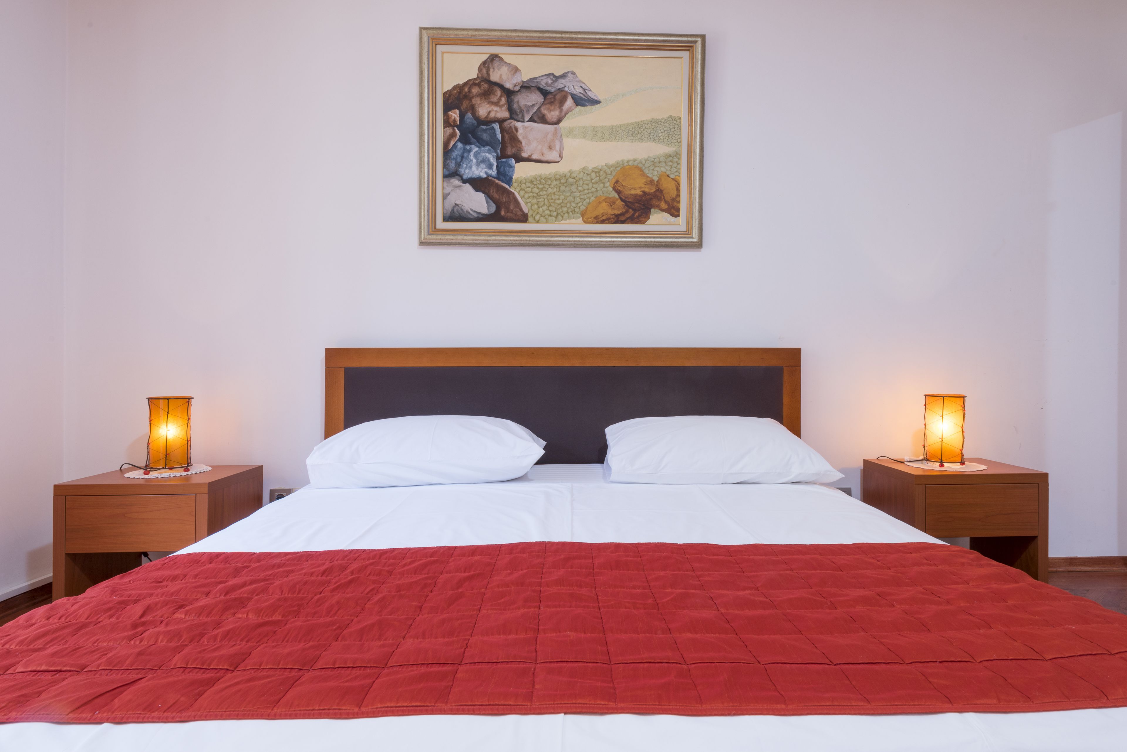 A neatly made bed with a red blanket, two white pillows, and a wooden headboard. There are two bedside tables with lamps, and a framed picture hanging on the wall.
