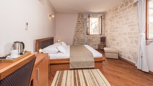 A bedroom with a wooden bed, a bedside table with a kettle, a tray with a cup and saucer, a small stool, and a stone wall. - Hotel Villa Nora (Hvar)