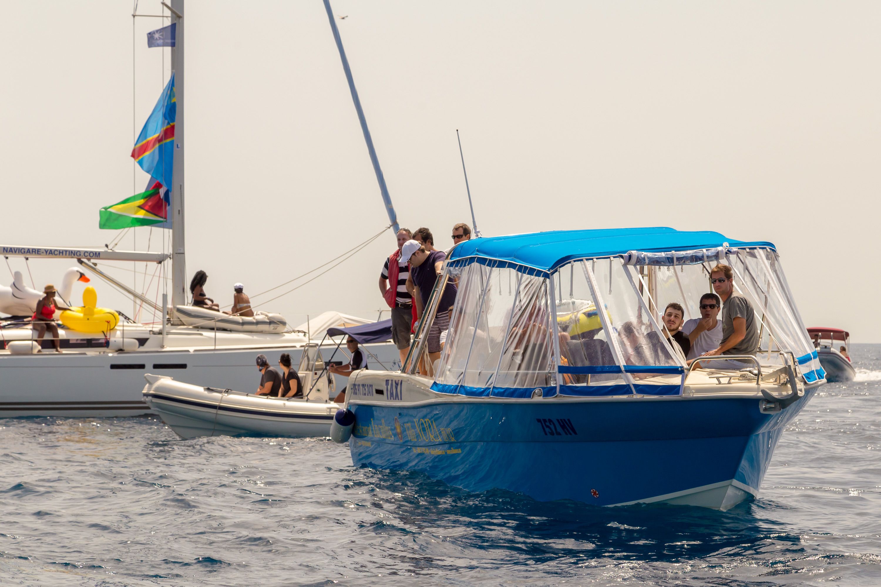 Un pequeño barco azul con toldo, personas a bordo y otro velero al fondo.