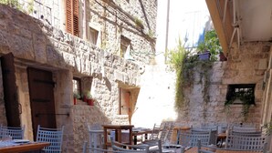 An outdoor dining area with stone walls, wooden tables, and white chairs. - Hotel Villa Nora (Hvar)