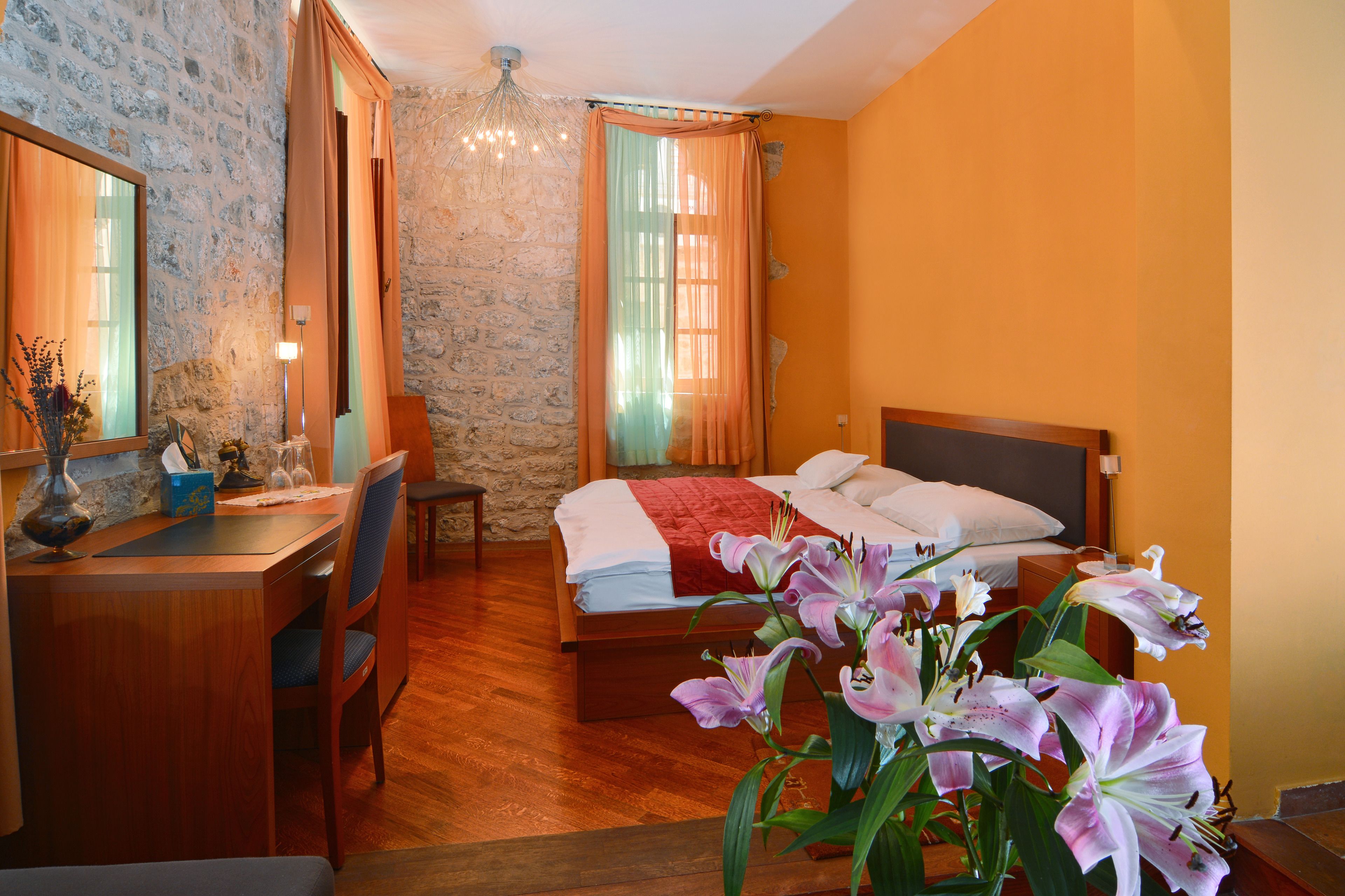 A hotel room with a stone wall, wooden furniture, a bed with a red blanket, and a vase of flowers.