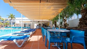 A poolside area with blue lounge chairs and tables, a covered seating area, and a building in the background. - BLUESEA Jandia Luz (Pajara)
