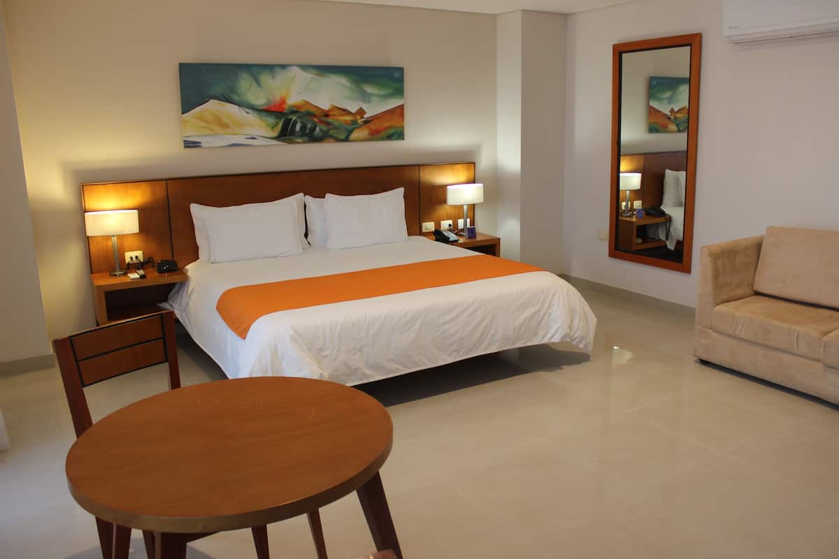 A hotel room with a bed, two bedside tables with lamps, a chair, a round table, and a painting on the wall.