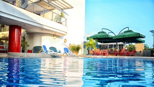 Una piscina con sillas de descanso y sombrillas, un pilar rojo y un edificio con balcones.