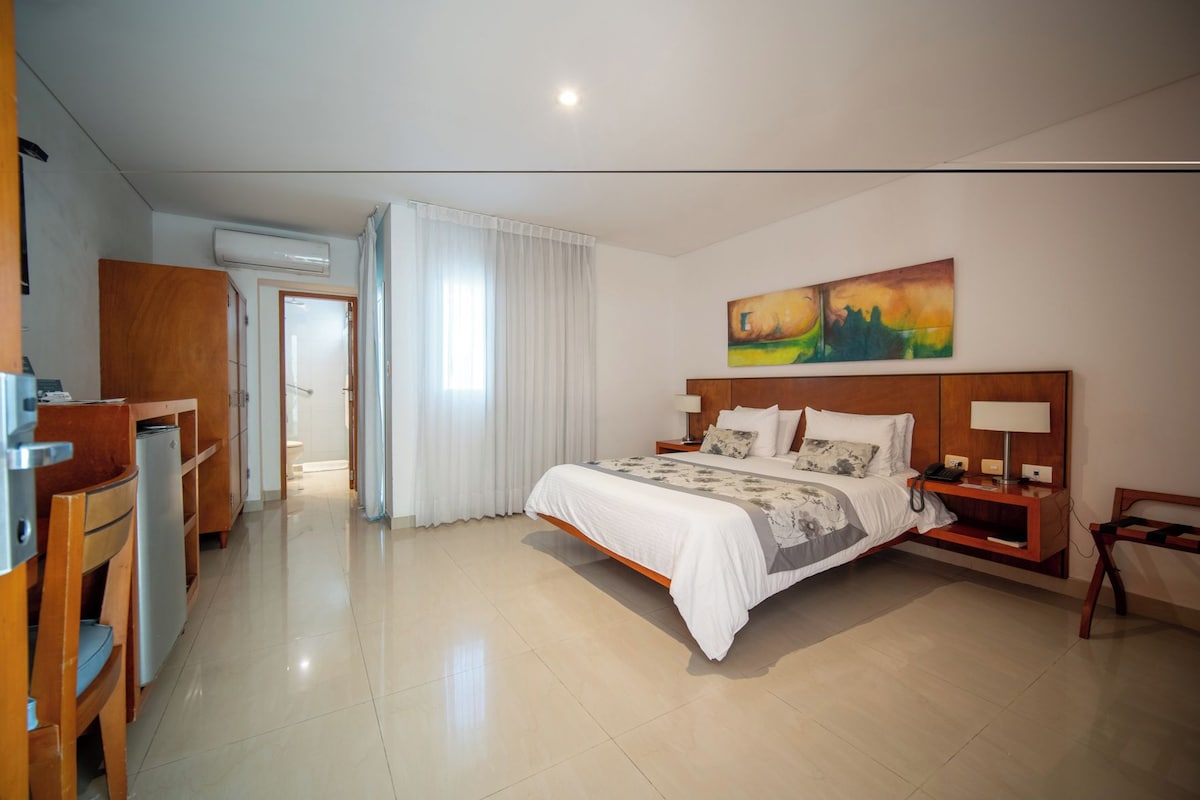 Habitación de hotel con una cama grande, un escritorio y una silla. Hay un cuadro en la pared y una ventana con cortinas.