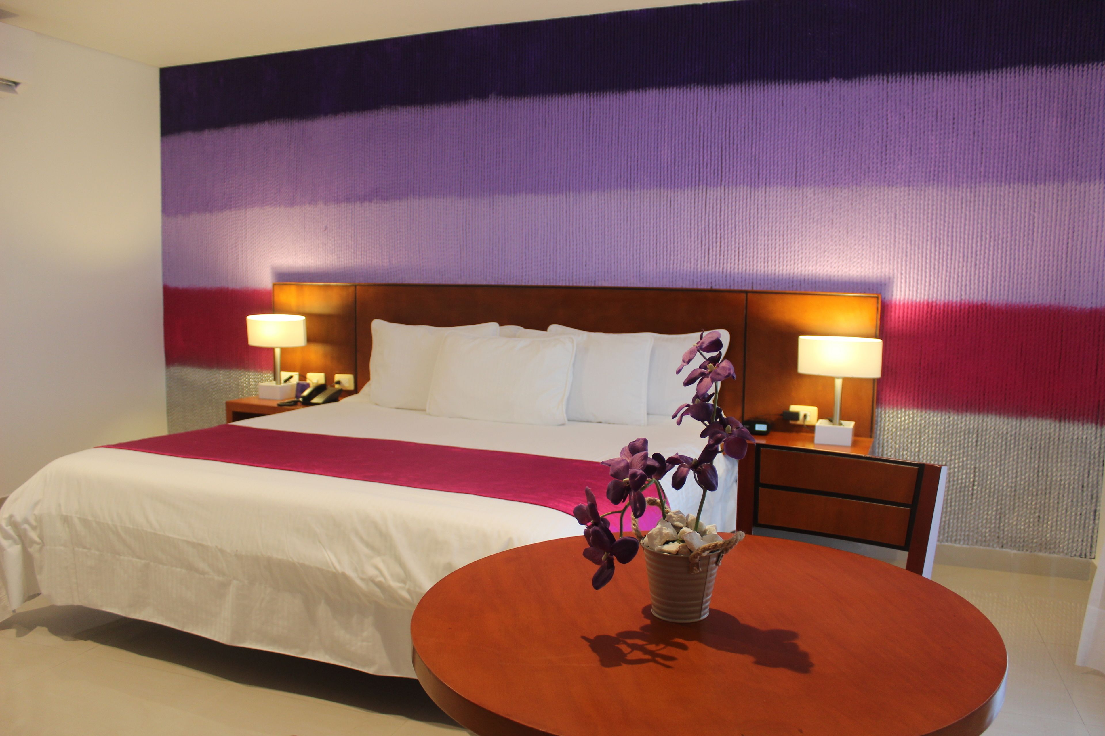 Habitación de hotel con una cama grande, dos mesitas de noche con lámparas, una mesa redonda de madera con un jarrón de flores y un mural de pared colorido.