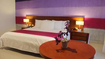 Habitación de hotel con una cama grande, dos mesitas de noche con lámparas, una mesa redonda de madera con un jarrón de flores y un mural de pared colorido.