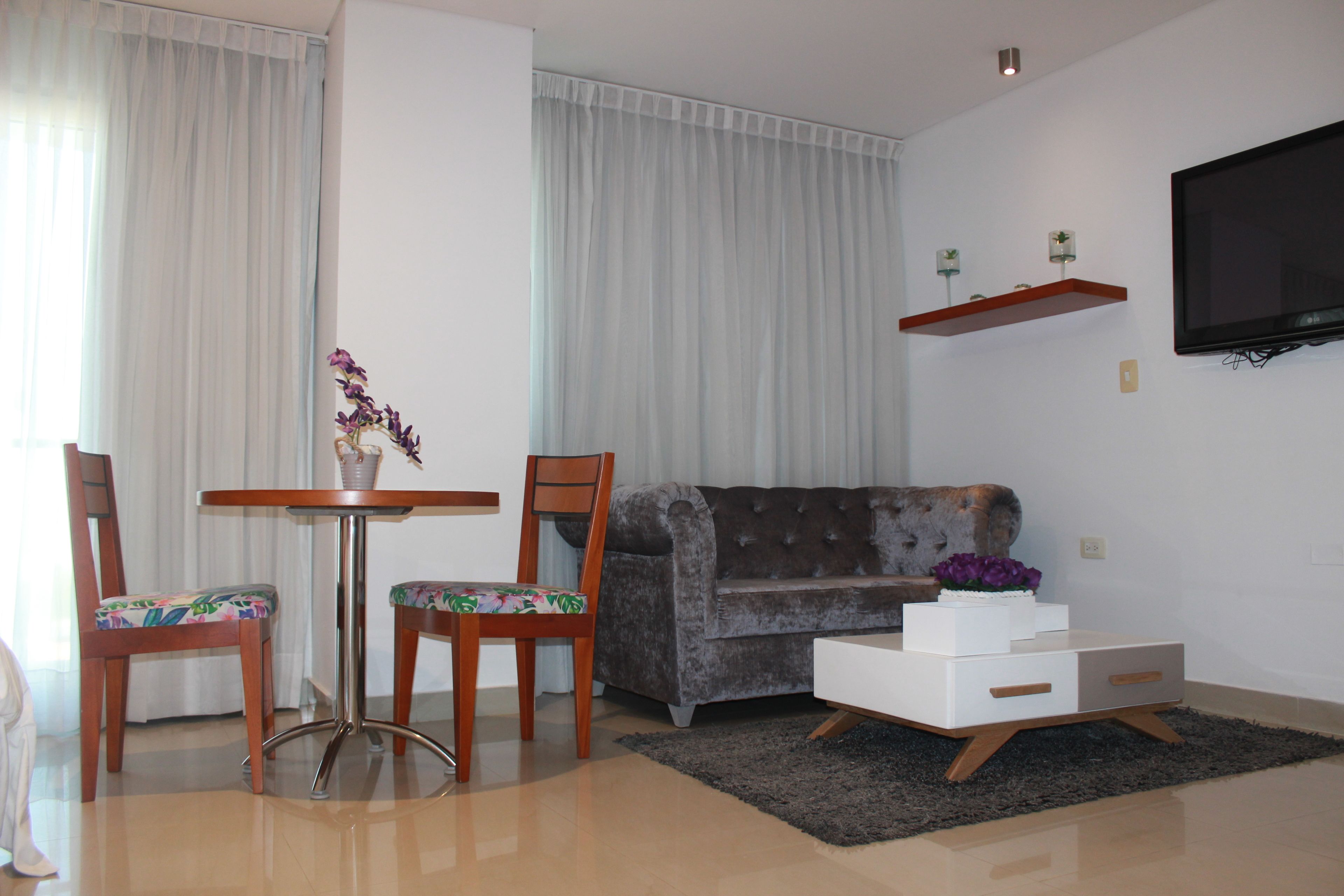 Una sala de estar con un sofá, dos sillones, una mesita, un televisor montado en la pared y una planta decorativa.