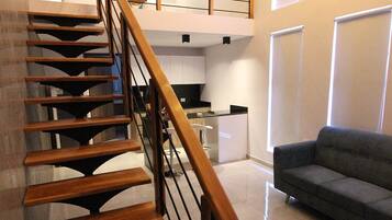 Un interior moderno con una escalera de madera que conduce a un dormitorio en entrepiso, una zona de cocina y una sala de estar con sofá.