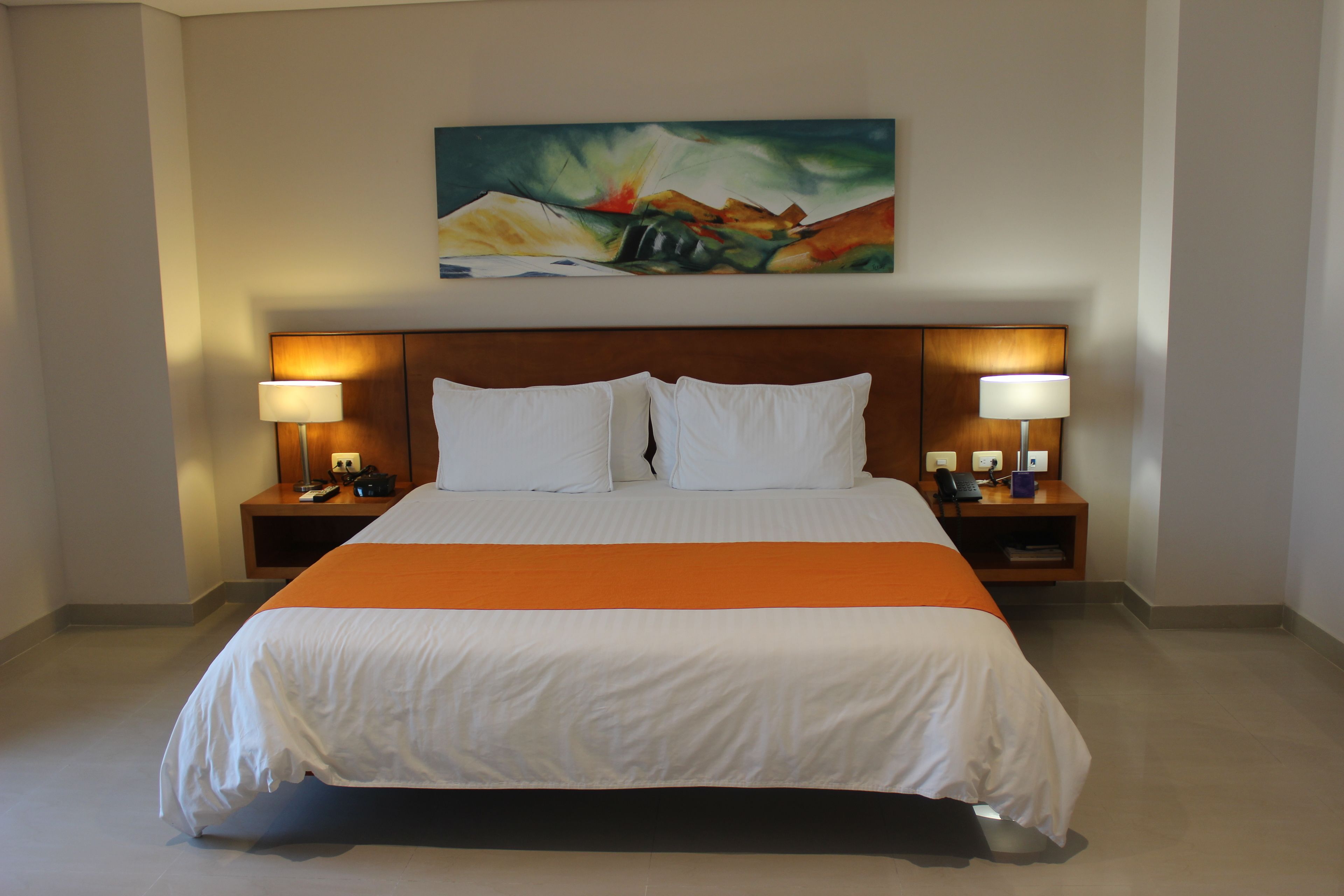 A hotel room with a large bed, two bedside tables with lamps, a painting on the wall, and a bright orange accent on the bedspread.