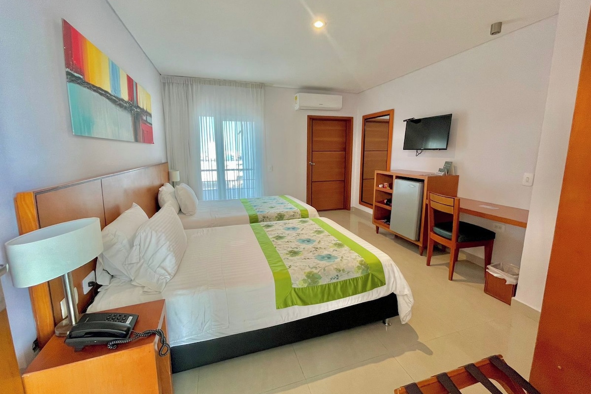 Habitación de hotel con cama, mesita de noche, lámpara, teléfono, televisión, escritorio, silla y ventana con cortinas.