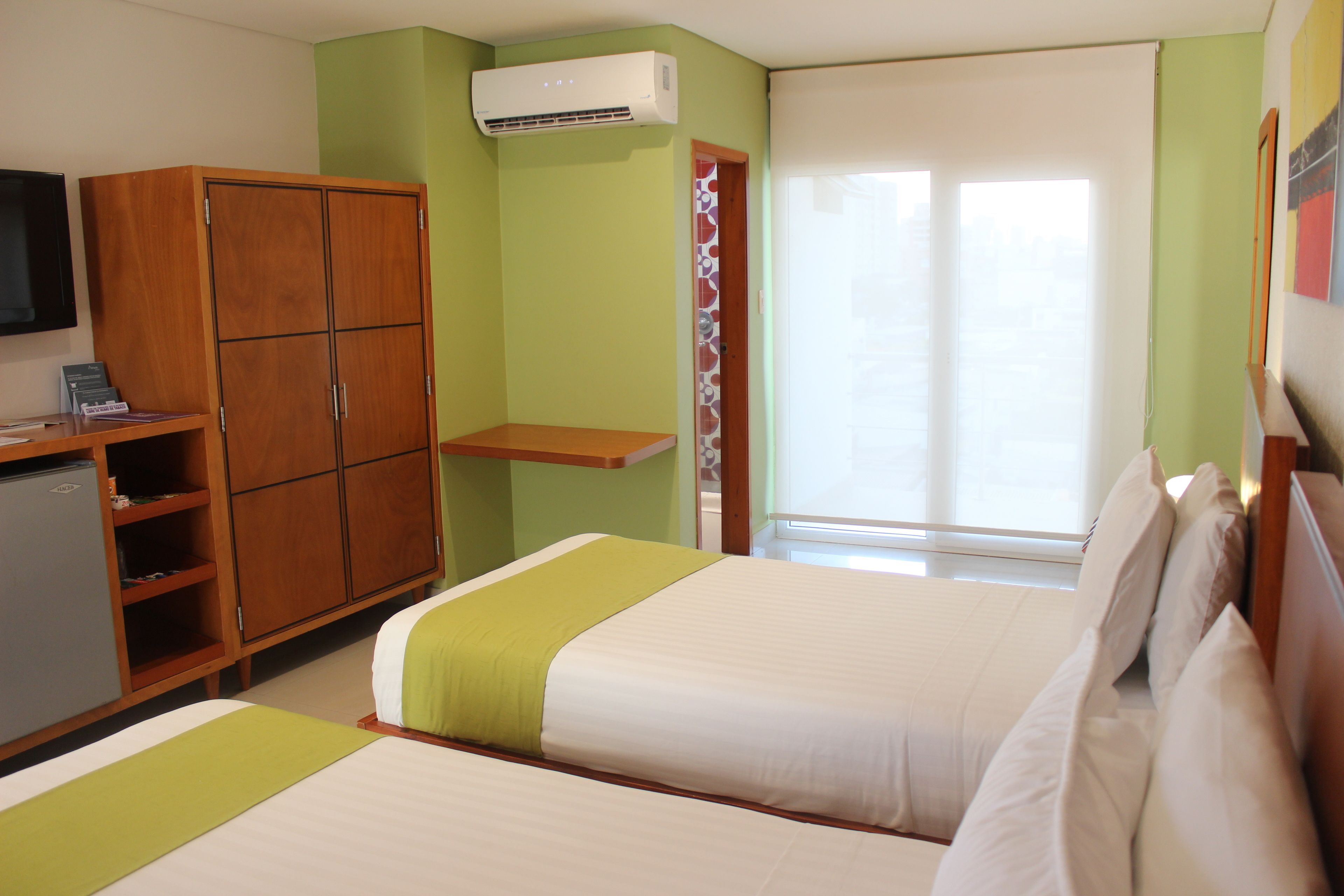 Habitación de hotel con dos camas, un armario, un escritorio con ordenador y una ventana con persianas.