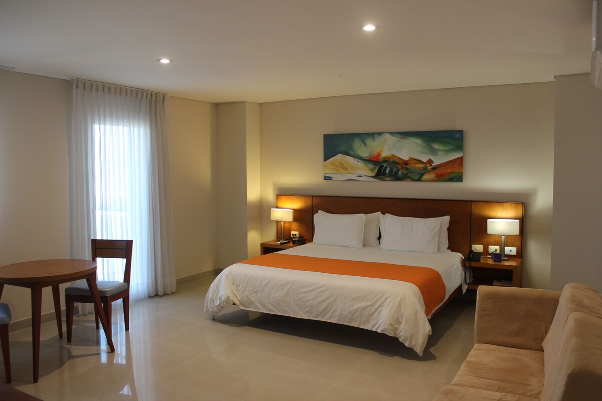 Habitación de hotel con cama, dos mesitas de noche con lámparas, una mesa de comedor con sillas y un cuadro en la pared.