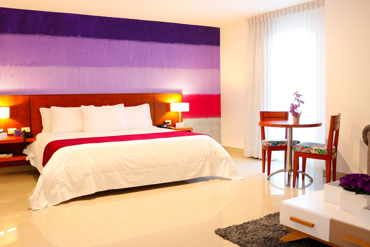 Habitación de hotel con una cama grande, un escritorio con silla, una mesita con un jarrón de flores y un sofá.