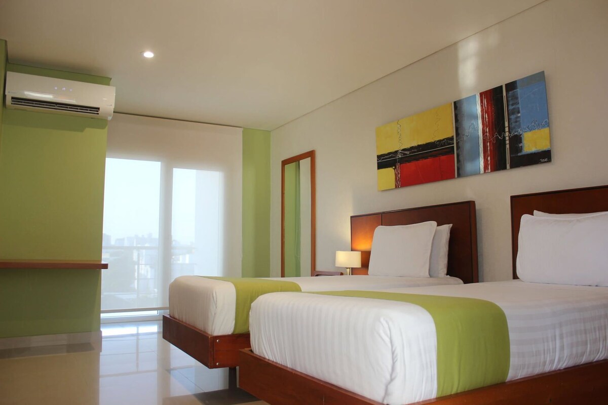 Una habitación de hotel con dos camas, un ventanal grande, un aire acondicionado de pared y un cuadro.