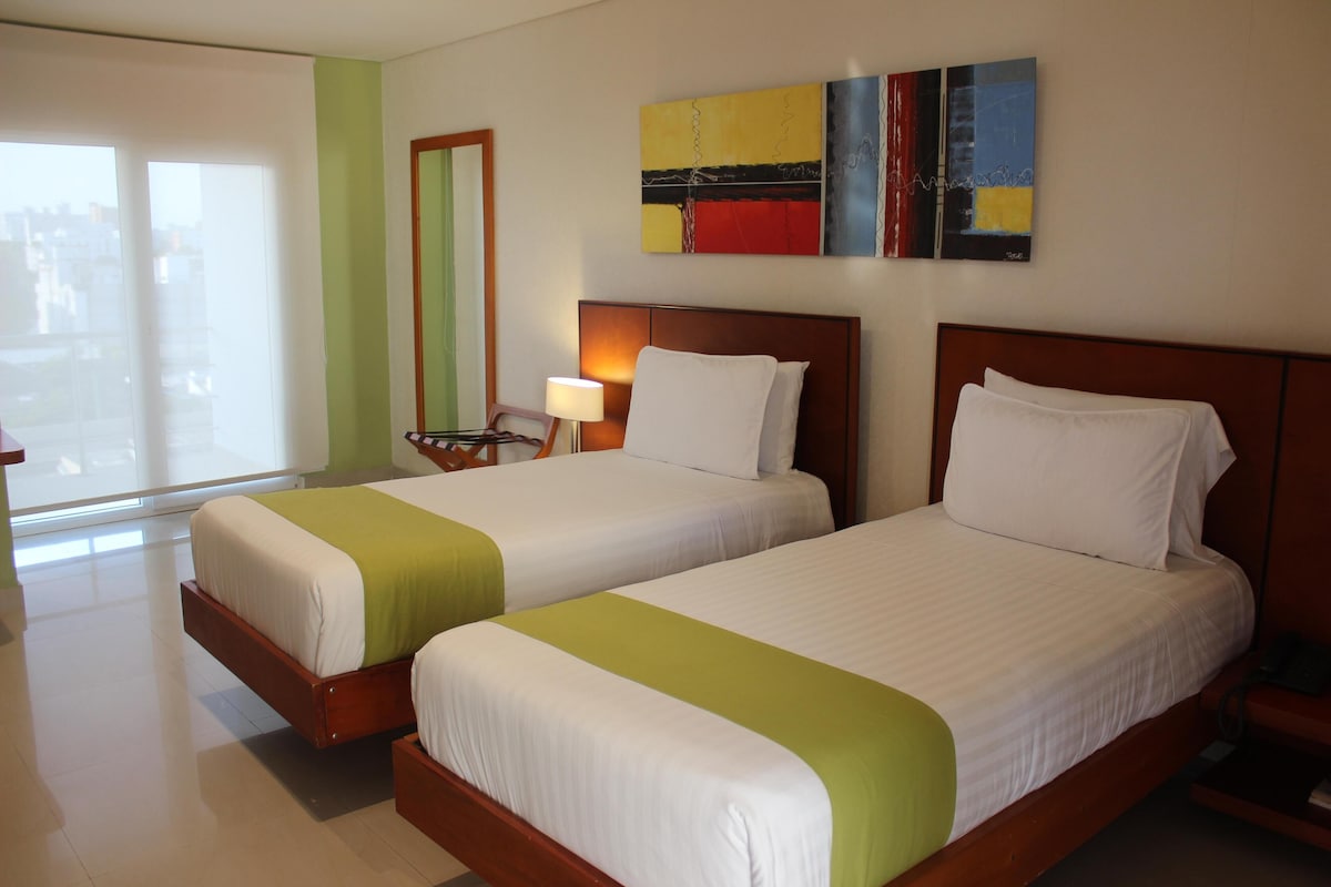 Un cuarto de hotel con dos camas, una mesita de noche con lámpara, un cuadro en la pared y vistas al exterior.