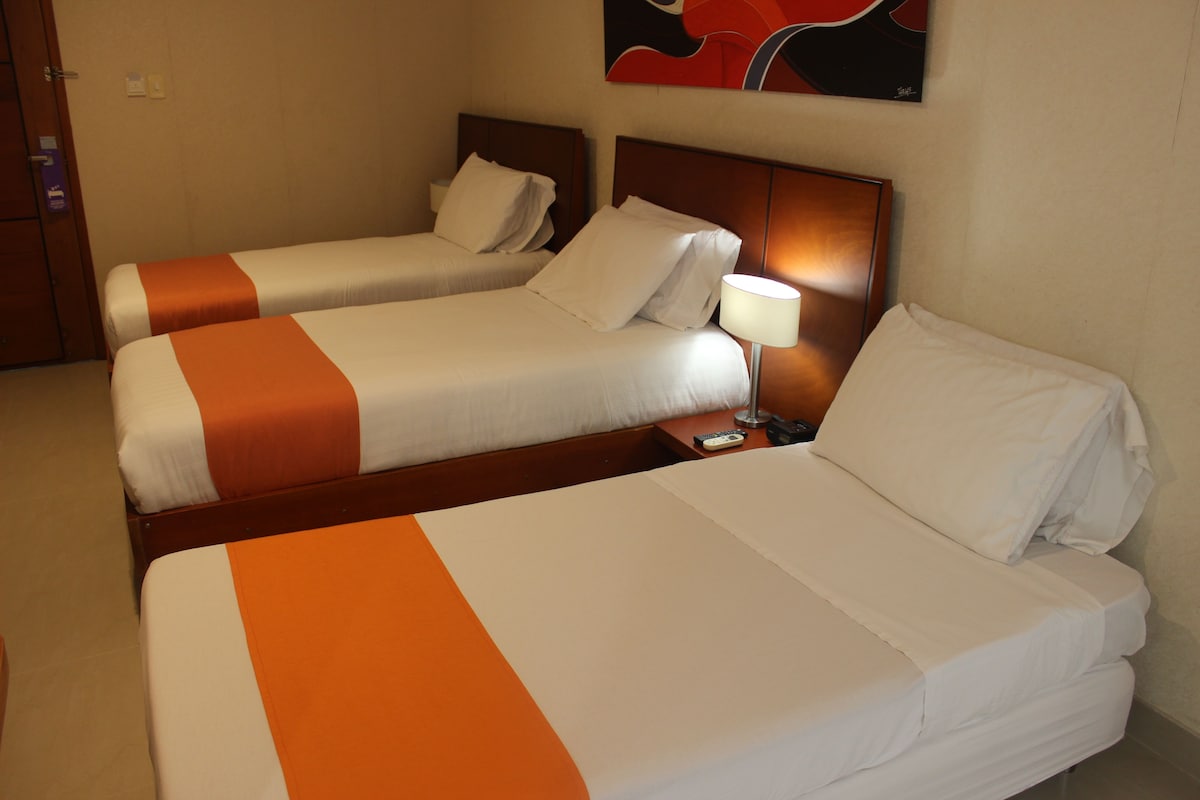 Habitación de hotel con dos camas, cada una con ropa blanca y detalles naranja, una mesita de noche con lámpara, y un cuadro en la pared.