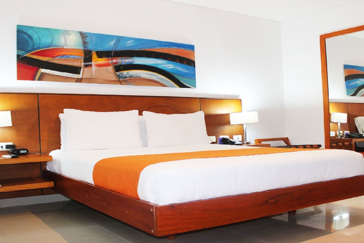 Habitación de hotel con una cama grande, dos mesitas de noche con lámparas, una mesita auxiliar, un espejo y un cuadro abstracto de gran tamaño en la pared.