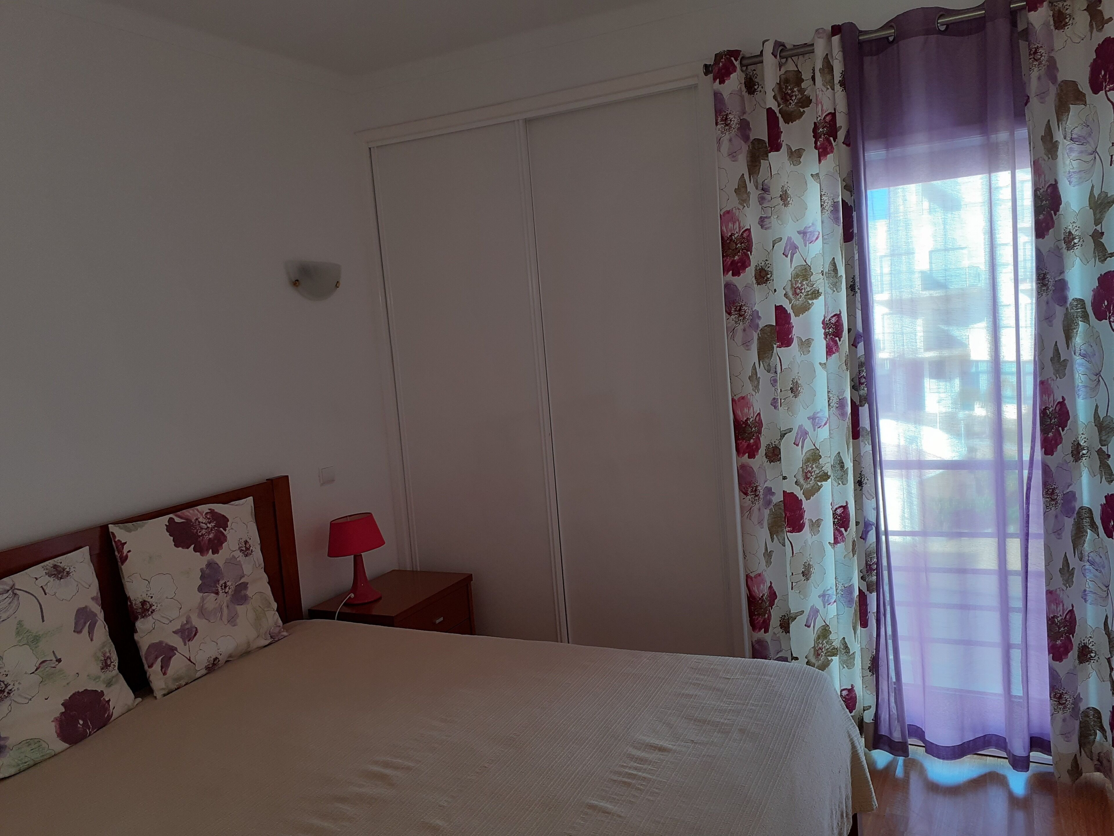 A bedroom with a bed, a nightstand with a lamp, a wardrobe, and floral curtains.