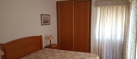 A bedroom with a bed, a nightstand, a wardrobe, a framed picture on the wall, and a window with curtains.