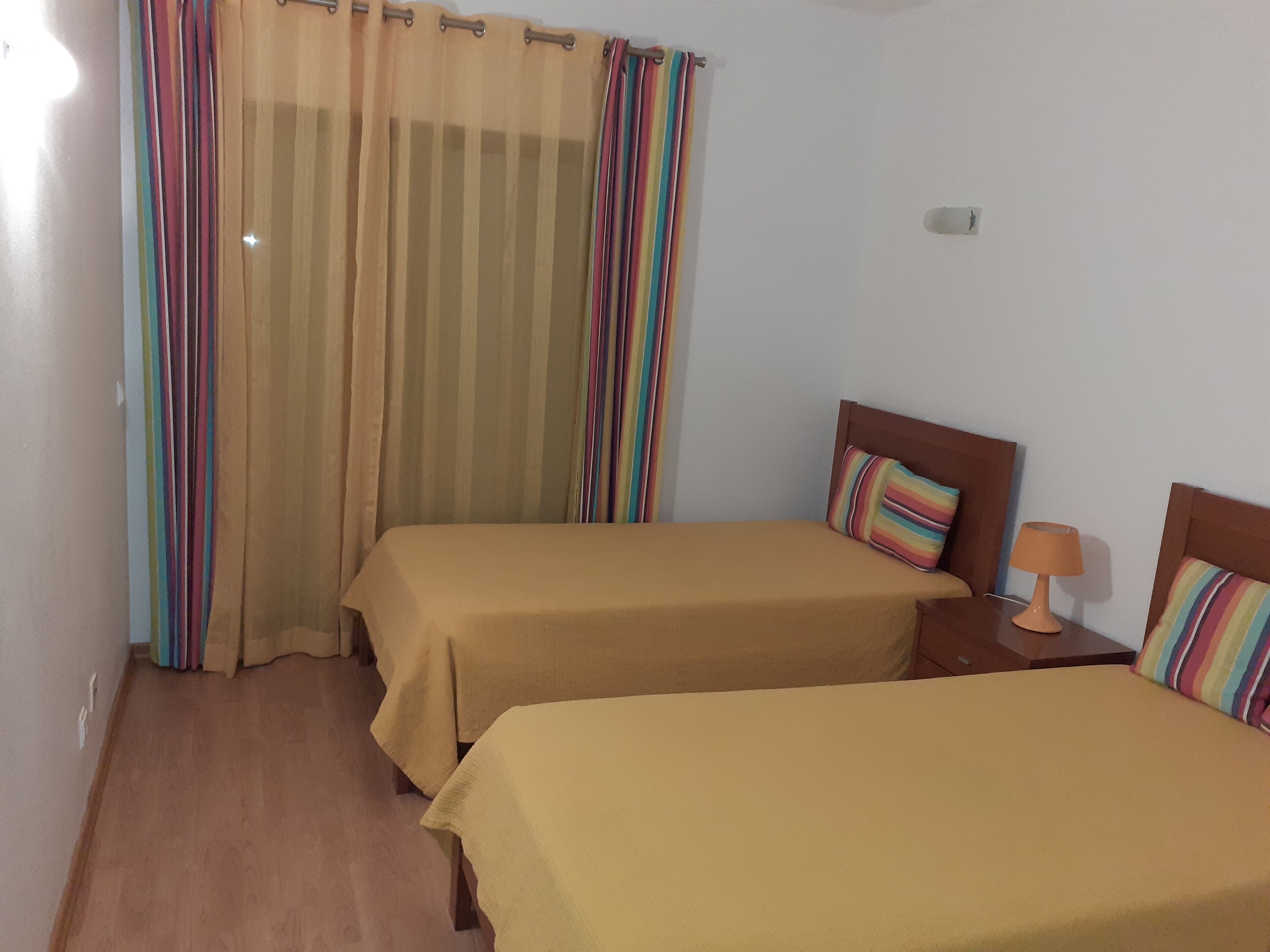 A room with two beds, each with a striped pillow, a small table with a lamp, and a curtain with multicolored stripes.
