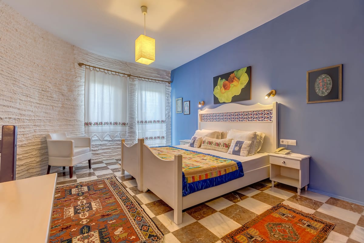 A bedroom with a checkered floor, a bed with a colorful quilt, a white chair, a nightstand with a phone, and a wall with framed pictures.