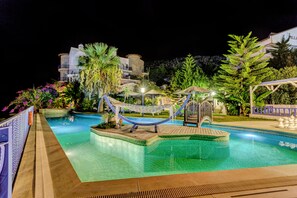 A nighttime swimming pool area with a hammock, lounge chairs, and a small bridge.