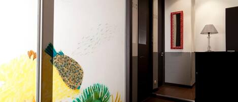 A hallway with a colorful wall mural, a red-framed mirror, and a lamp on a console table.