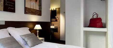 A hotel room with a bed, pillows, a nightstand, a lamp, a red handbag on a shelf, and artwork on the wall.