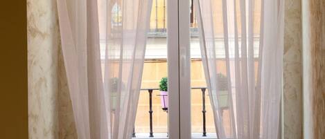 Une chambre avec un lit, une fenêtre ornée de rideaux légers et donnant sur le balcon extérieur.