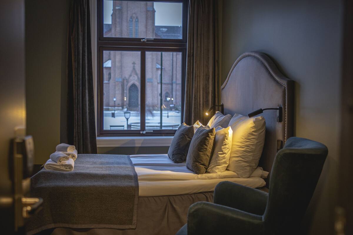 Habitación de hotel con cama, una silla, un ventanal con vista a un edificio y una mesa con toallas.