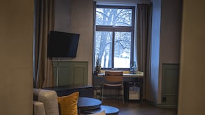 A living room with a sofa, a TV mounted on the wall, a dining table with chairs, and a window with a view of trees.