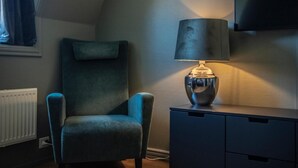 A room with a dark blue armchair, a lamp on a dresser, and a television on the wall.