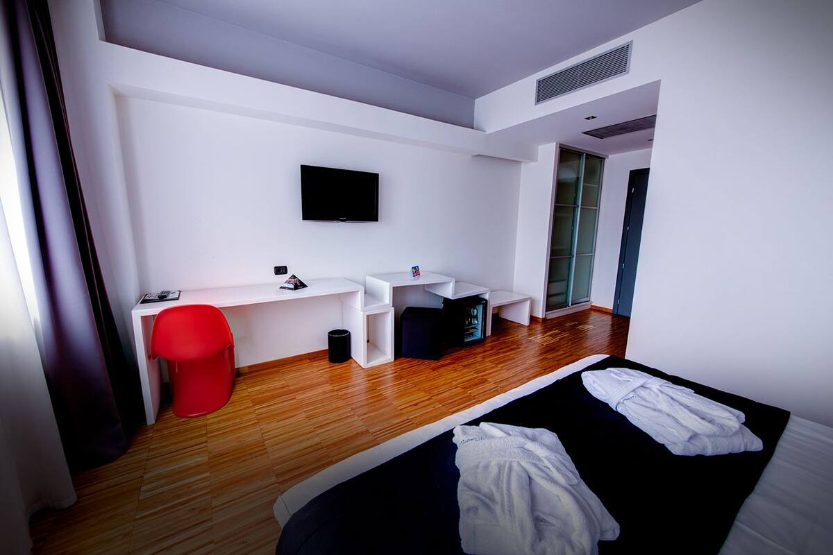 A modern hotel room with a wooden floor, a bed with white linens, a red chair, a desk with a flat-screen TV, and a small trash bin.