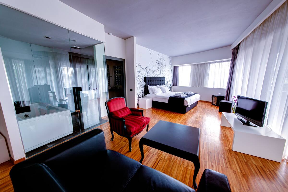 A modern hotel room with a bed, a TV, a red armchair, a sofa, and a glass partition separating the living area from the bedroom.