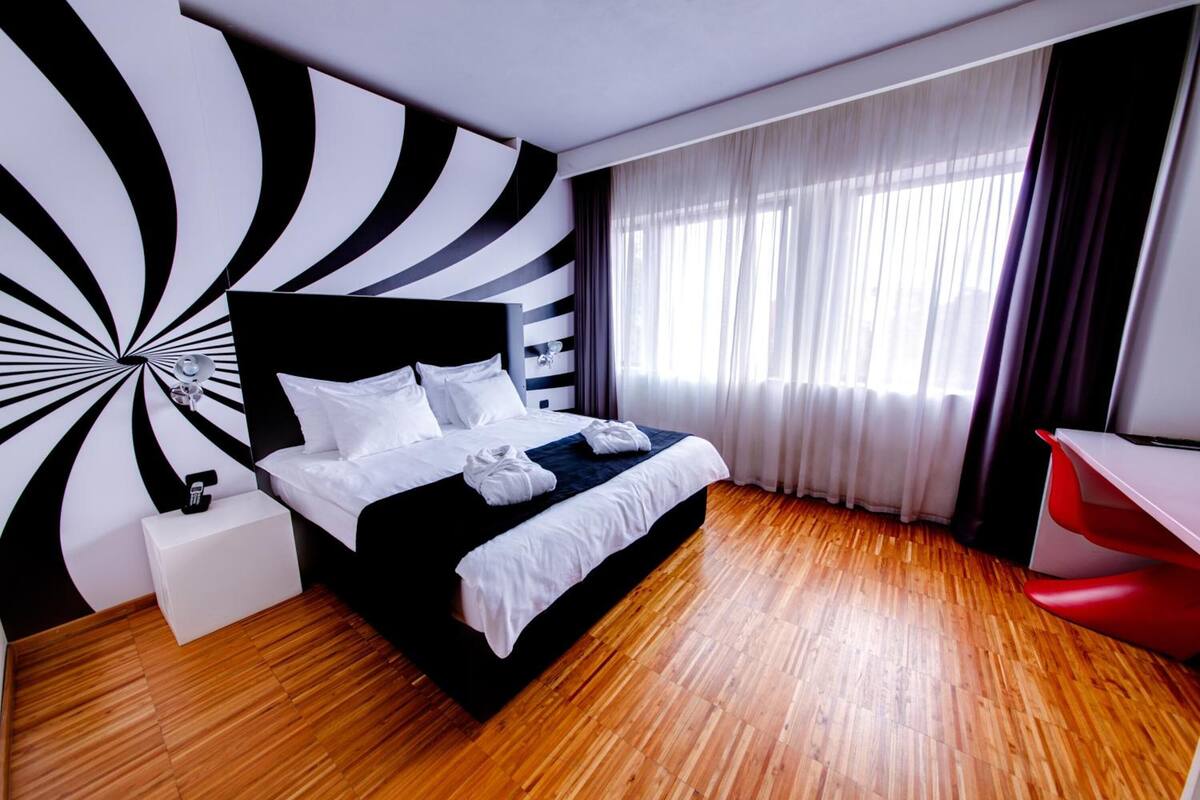 A modern hotel room with a black and white striped wall design, a wooden floor, a bed with white linens, a red desk, and a large window with sheer curtains.