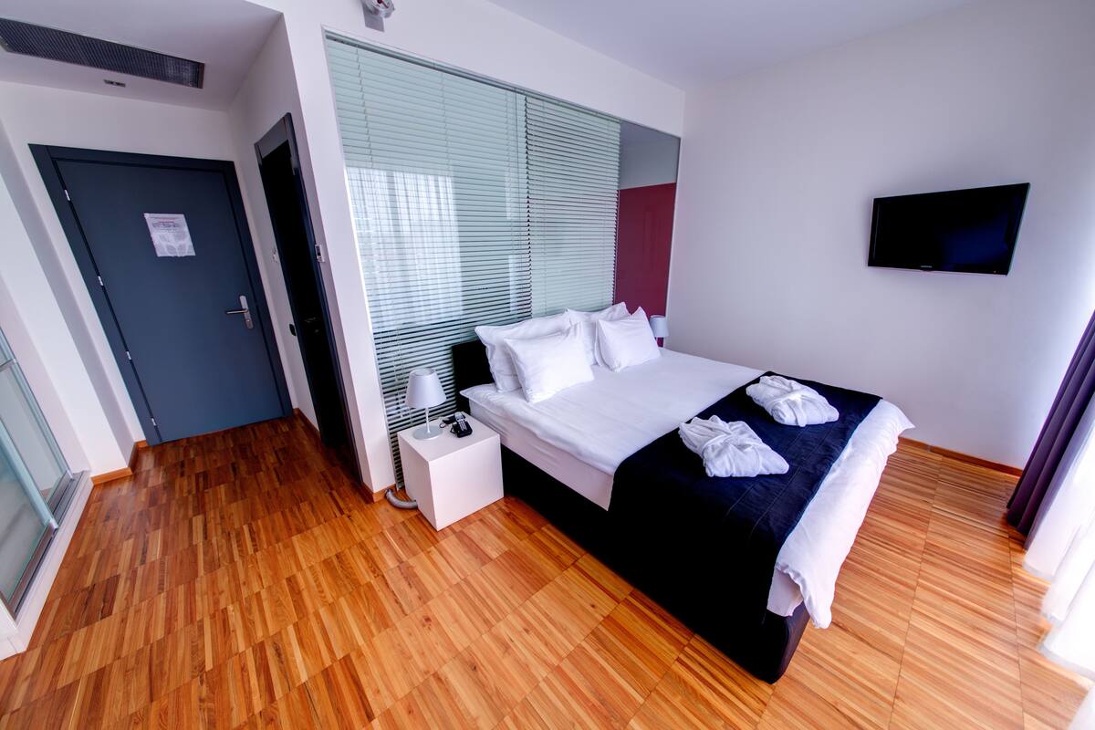 A modern hotel room with a large bed, a flat-screen TV mounted on the wall, and a wooden floor.