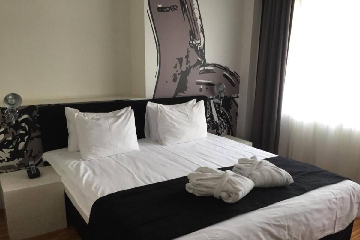 A hotel room with a bed, white pillows, a black headboard, a nightstand with a phone, and a wall with a graphic design.